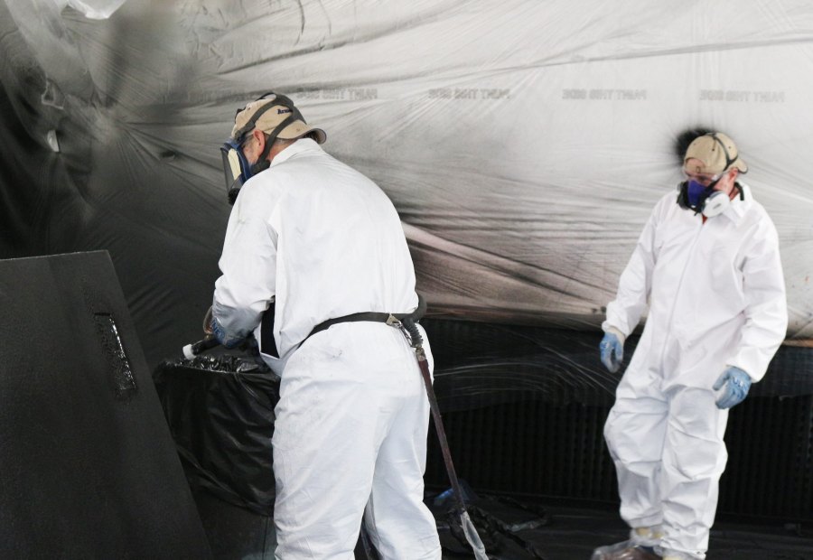 Inside a draped spray booth, an applicator sprays a dark panel while a second trainee observes