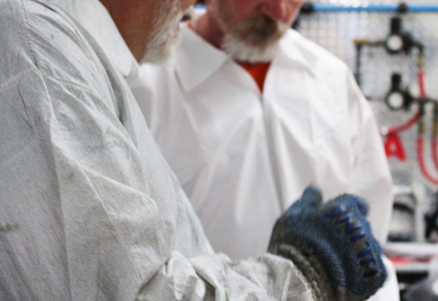 Instructor handing a spray gun to a trainee, both in protective suits