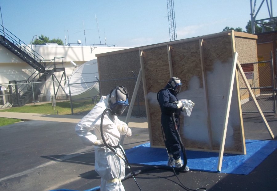 Two trainees in full protective gear spraying polyurea onto a framed panel during an outdoor Coatings Academy class