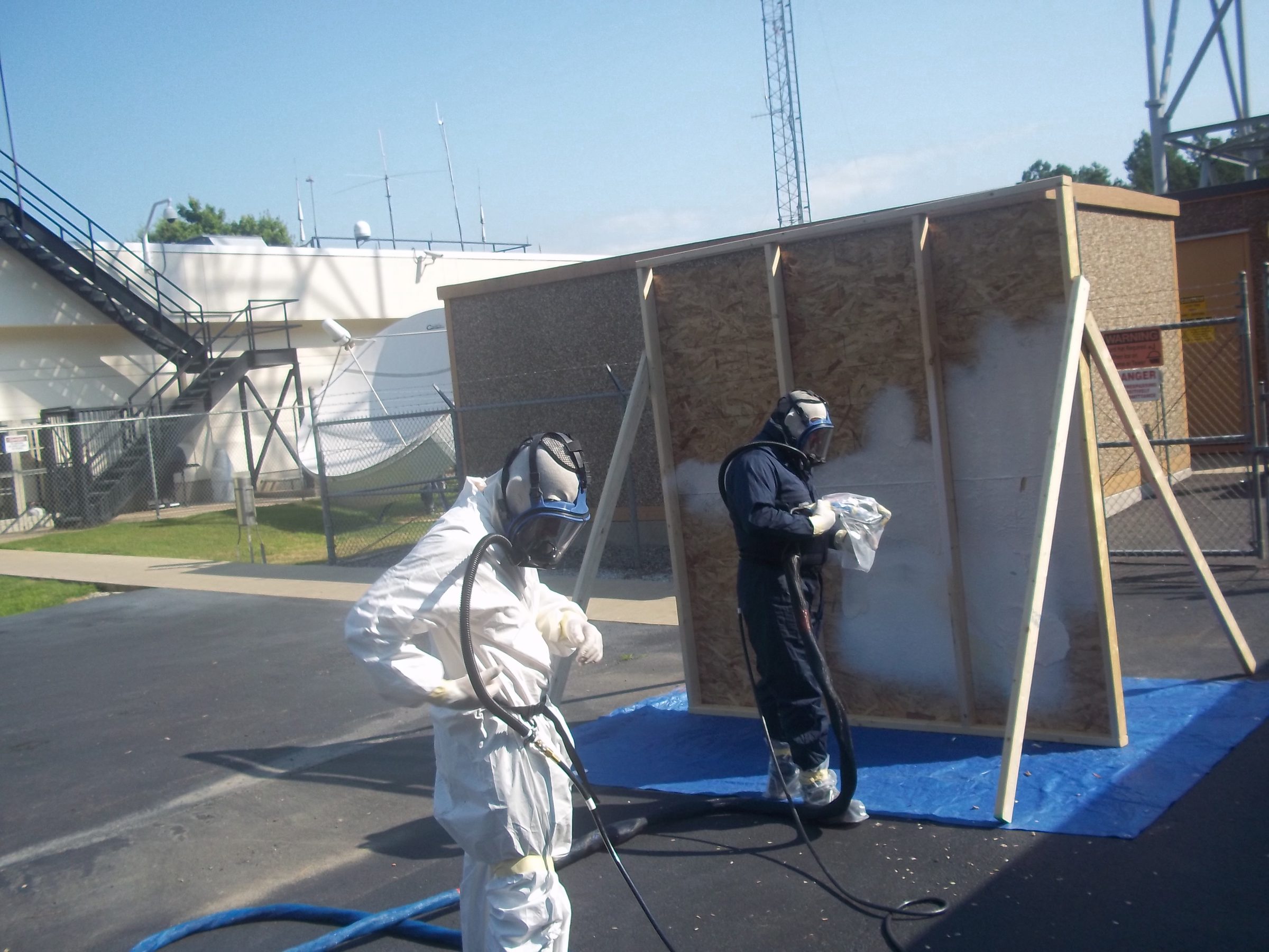 Two trainees in full protective gear spraying polyurea onto a framed panel during an outdoor Coatings Academy class