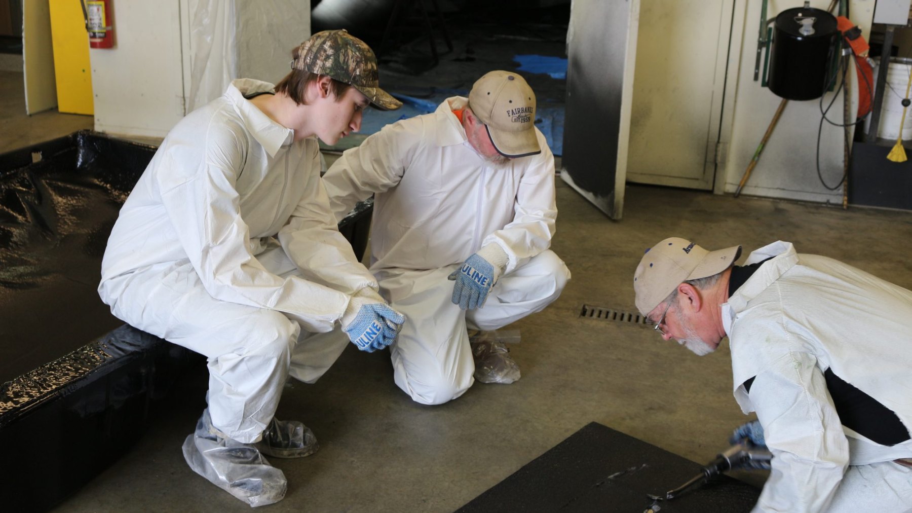 Three trainees kneeling on a shop floor inspecting the edge of a sprayed panel