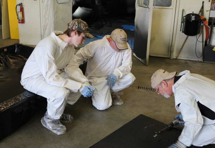 Three trainees kneeling on a shop floor inspecting the edge of a sprayed panel