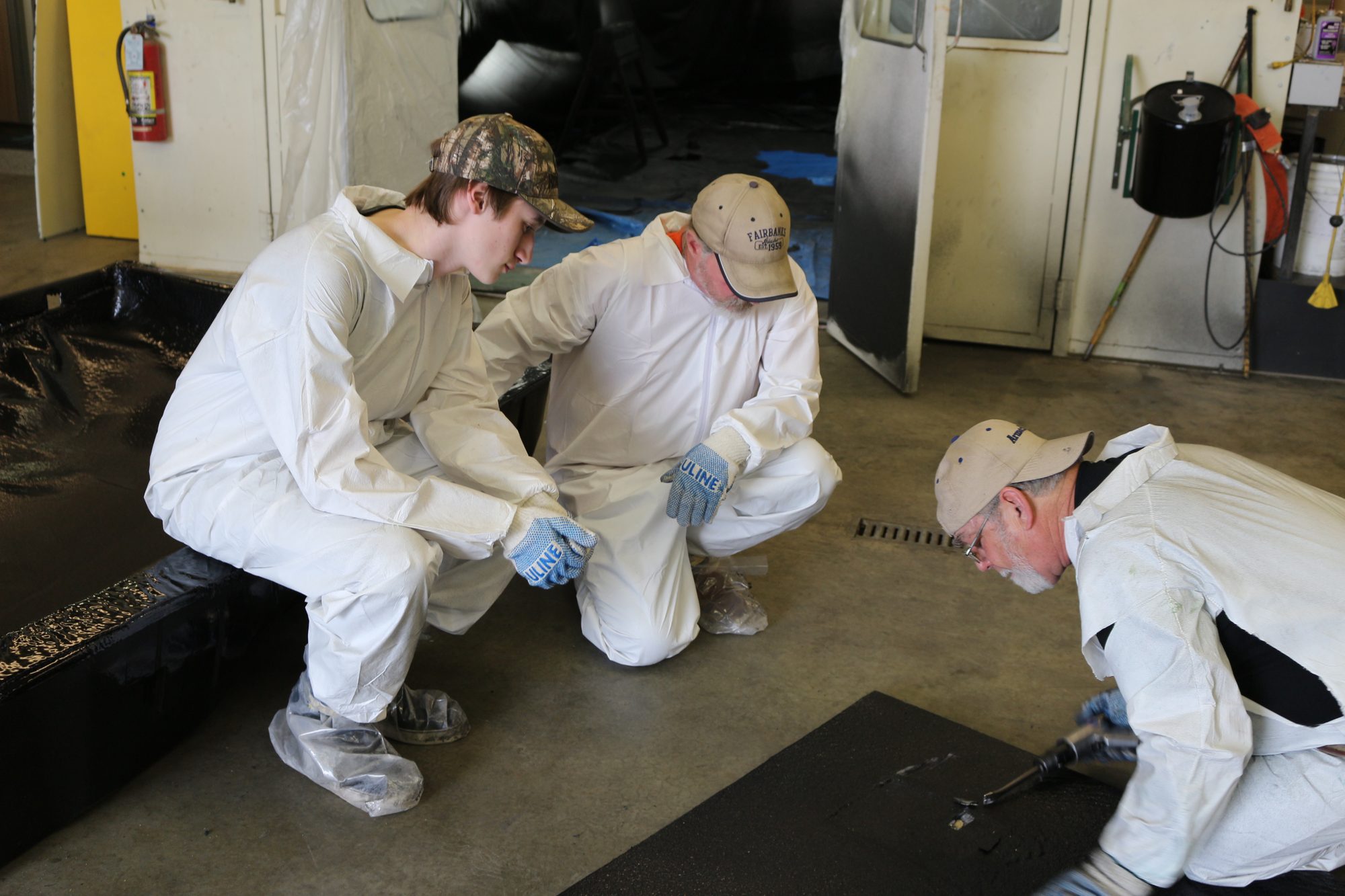 Three trainees kneeling on a shop floor inspecting the edge of a sprayed panel