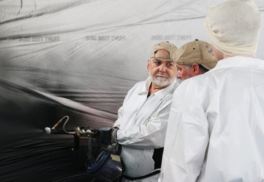 Instructor holding a heated spray gun and whip hose, turning to coach a trainee