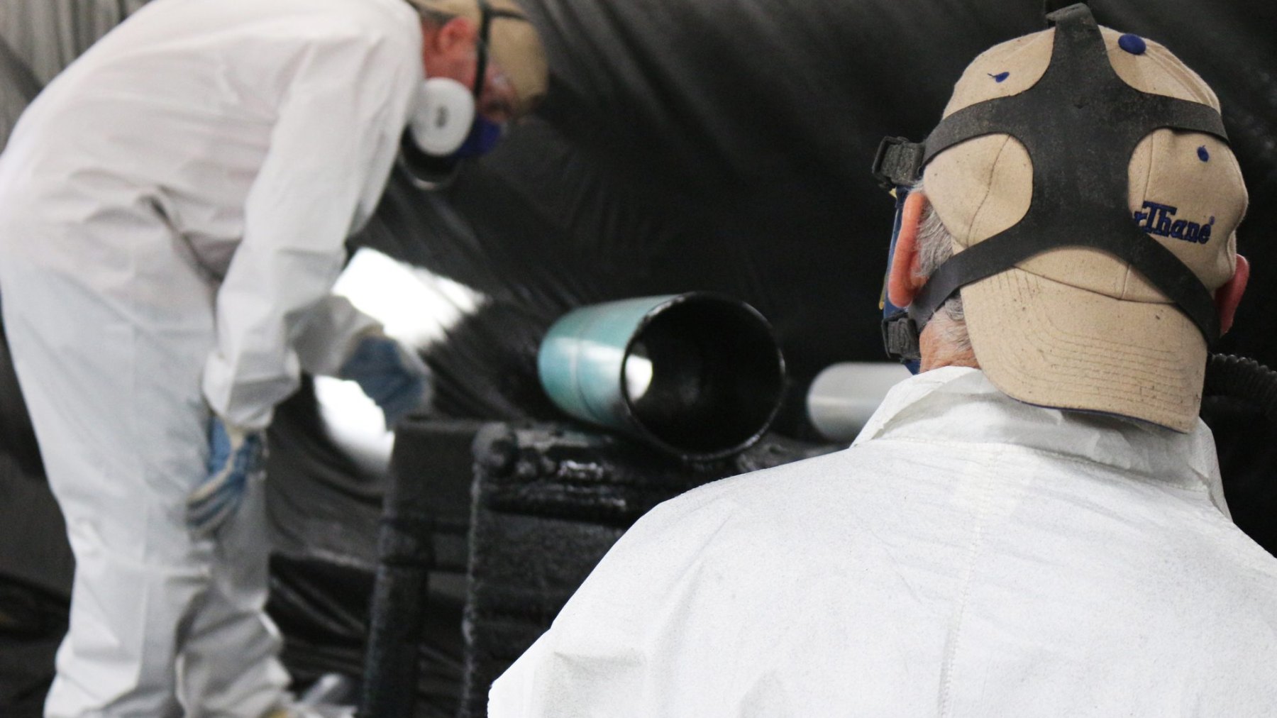 Two applicators in full-face respirators working on a pipe-lining setup inside a booth