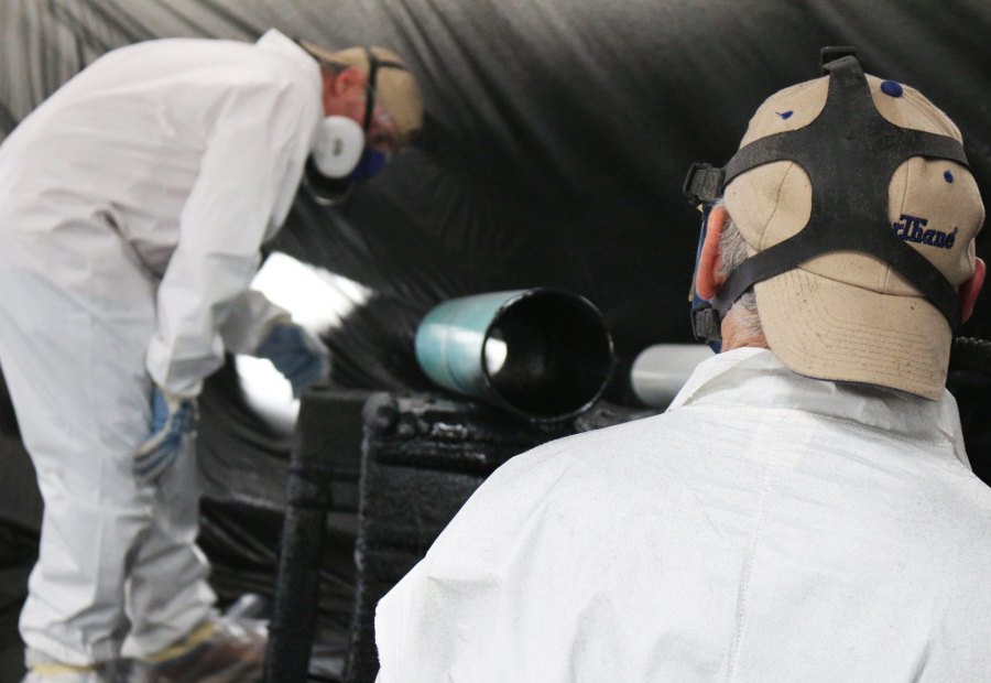 Two applicators in full-face respirators working on a pipe-lining setup inside a booth